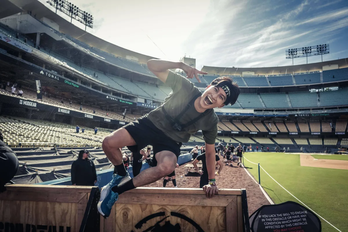 Maxwell Yeo grinning as he vaults a wooden obstacle wall in a stadium, arm cocked mid-swing