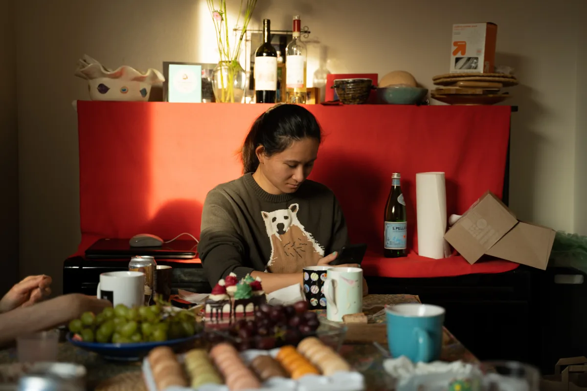 A woman looks at her phone at a table crowded with fruit, cakes and mugs, a red backdrop behind her