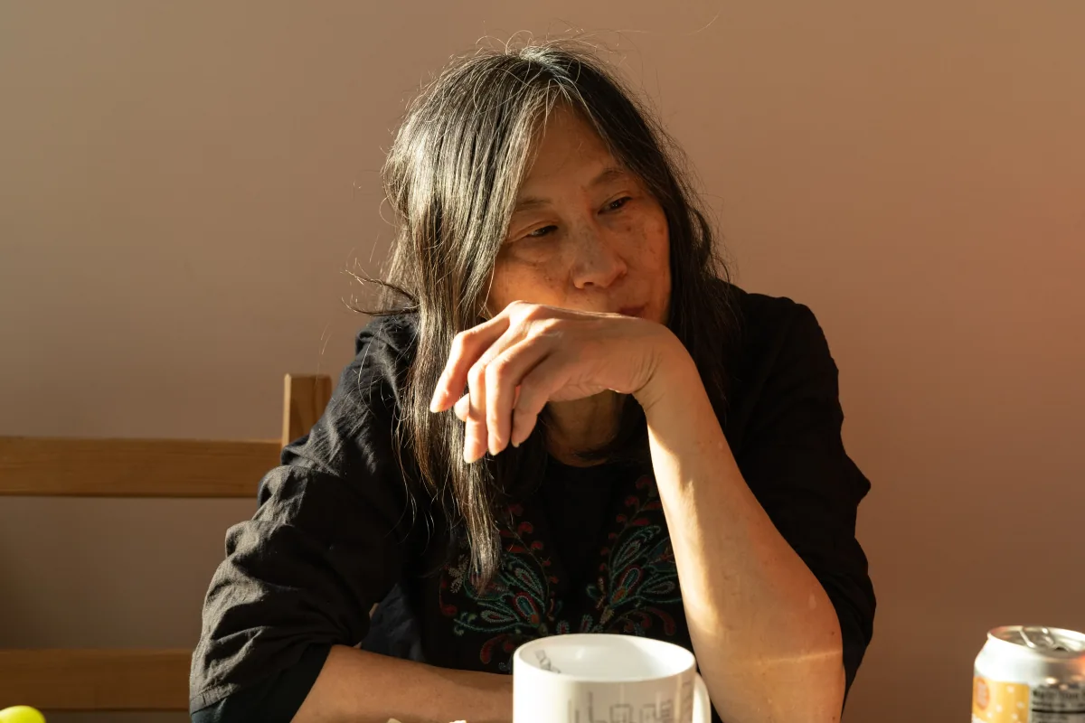 An older woman rests her chin on her hand at a table, warm afternoon light across her face