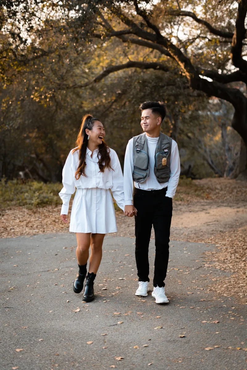 A couple in white walk hand in hand down a park path, laughing at each other under oak branches