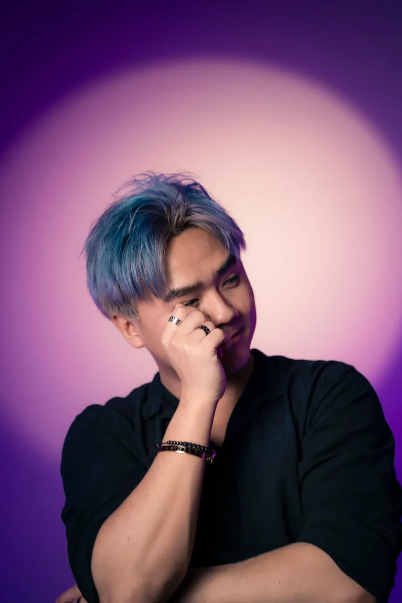 Studio portrait against a purple backdrop, blue-dyed hair, one hand covering an eye mid-laugh