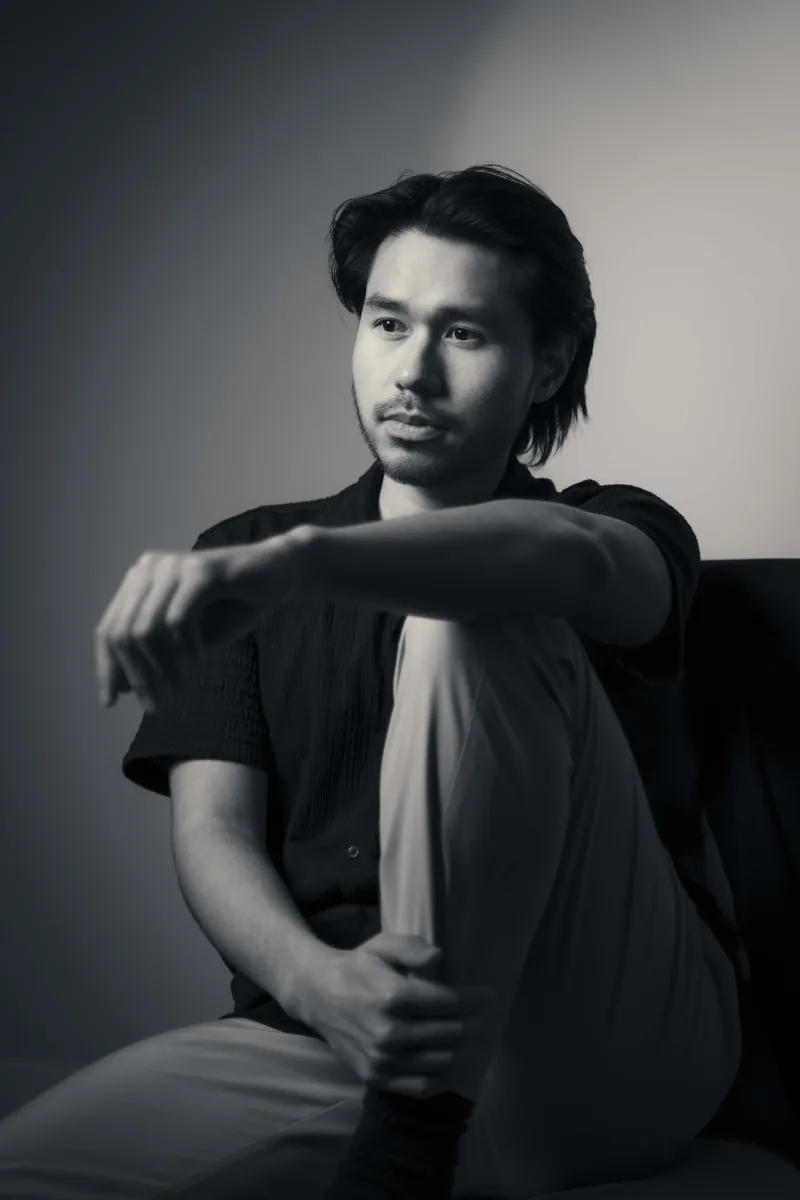 Black and white studio portrait of a man seated with one knee drawn up, arm draped over it
