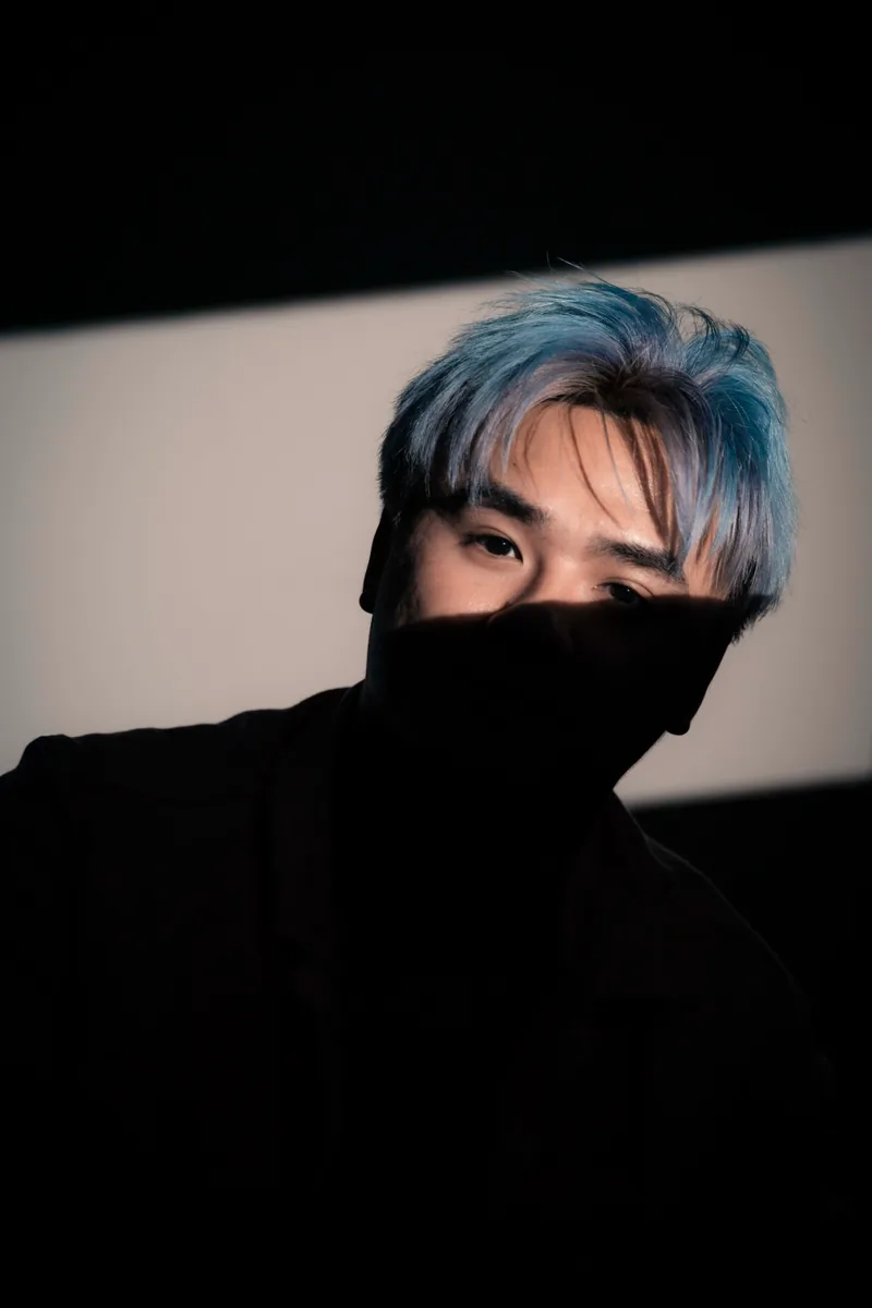 Blue-haired figure half-swallowed by shadow, only the eyes lit by a hard band of light on the wall behind