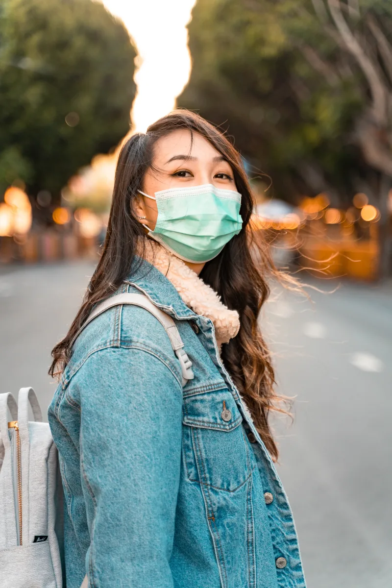 A woman in a denim jacket and surgical mask turns to the camera on an empty street at golden hour