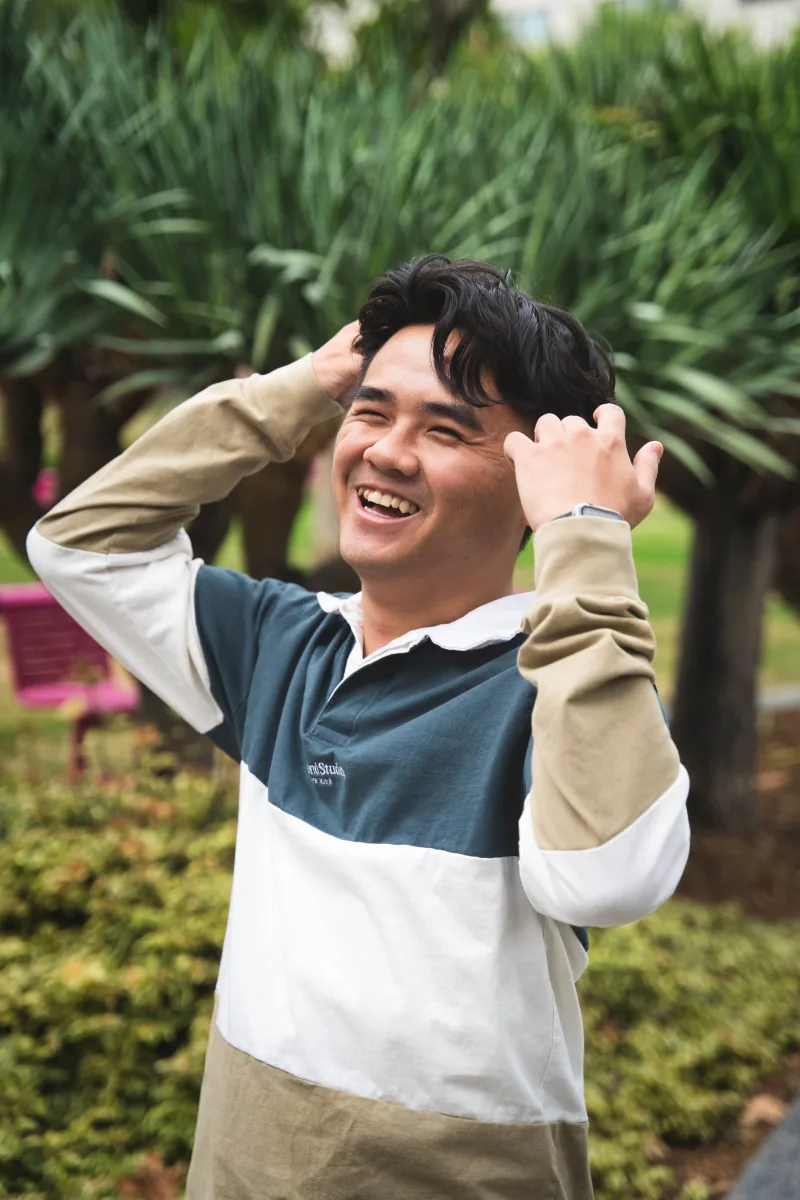 A man laughs with both hands in his hair, spiky green foliage behind him