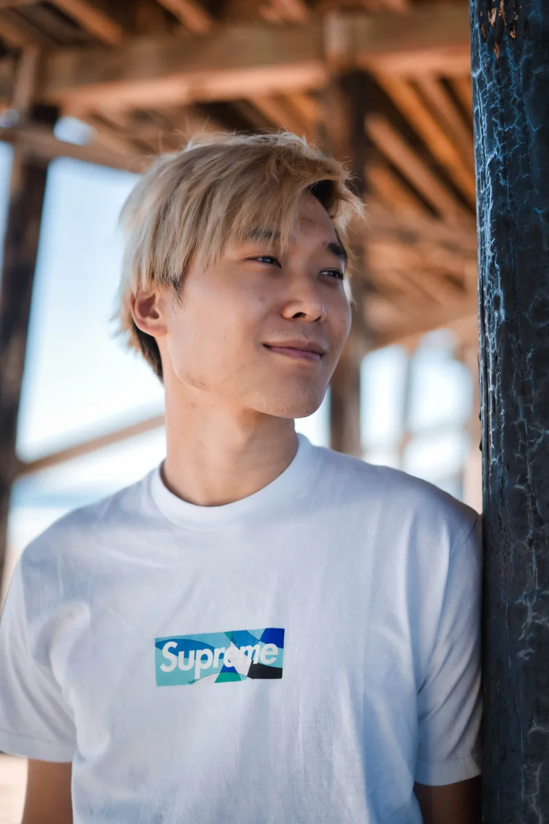 A man with bleached hair leans against a piling under a pier, half-smiling into the light