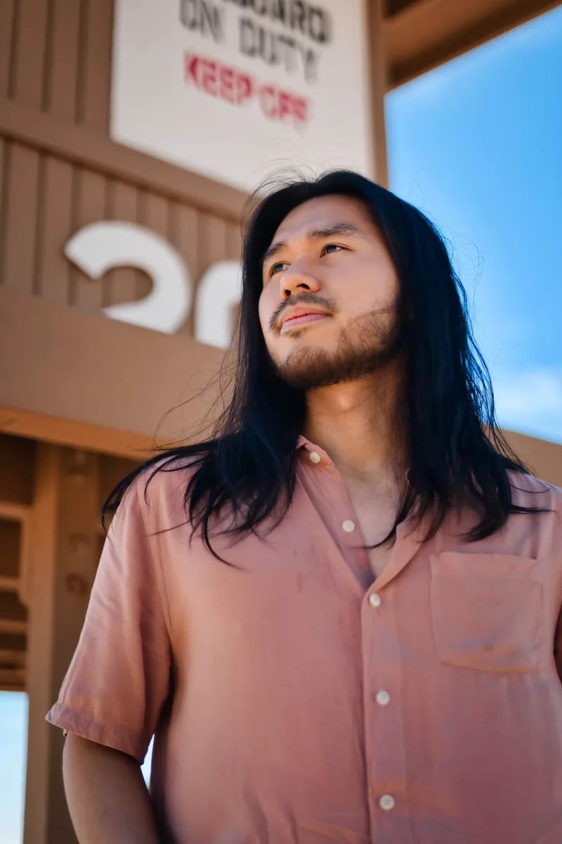 Low-angle portrait of a man with long dark hair in a pink shirt, a lifeguard tower behind him