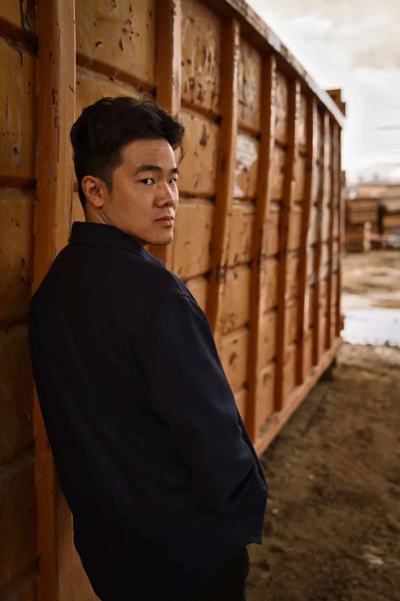 A man in a dark jacket leans against a rust-orange shipping container, looking back over his shoulder