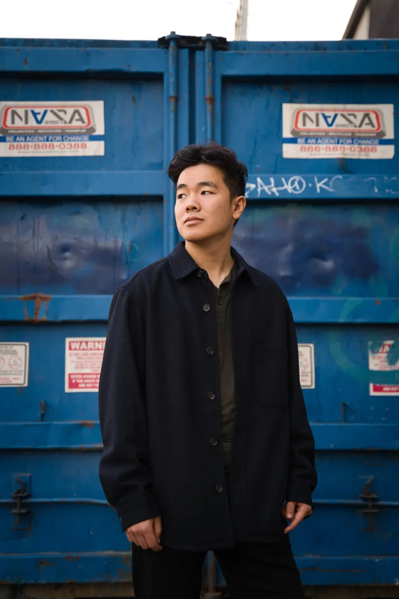 A man in a navy overshirt stands in front of a graffitied blue dumpster, looking off to one side