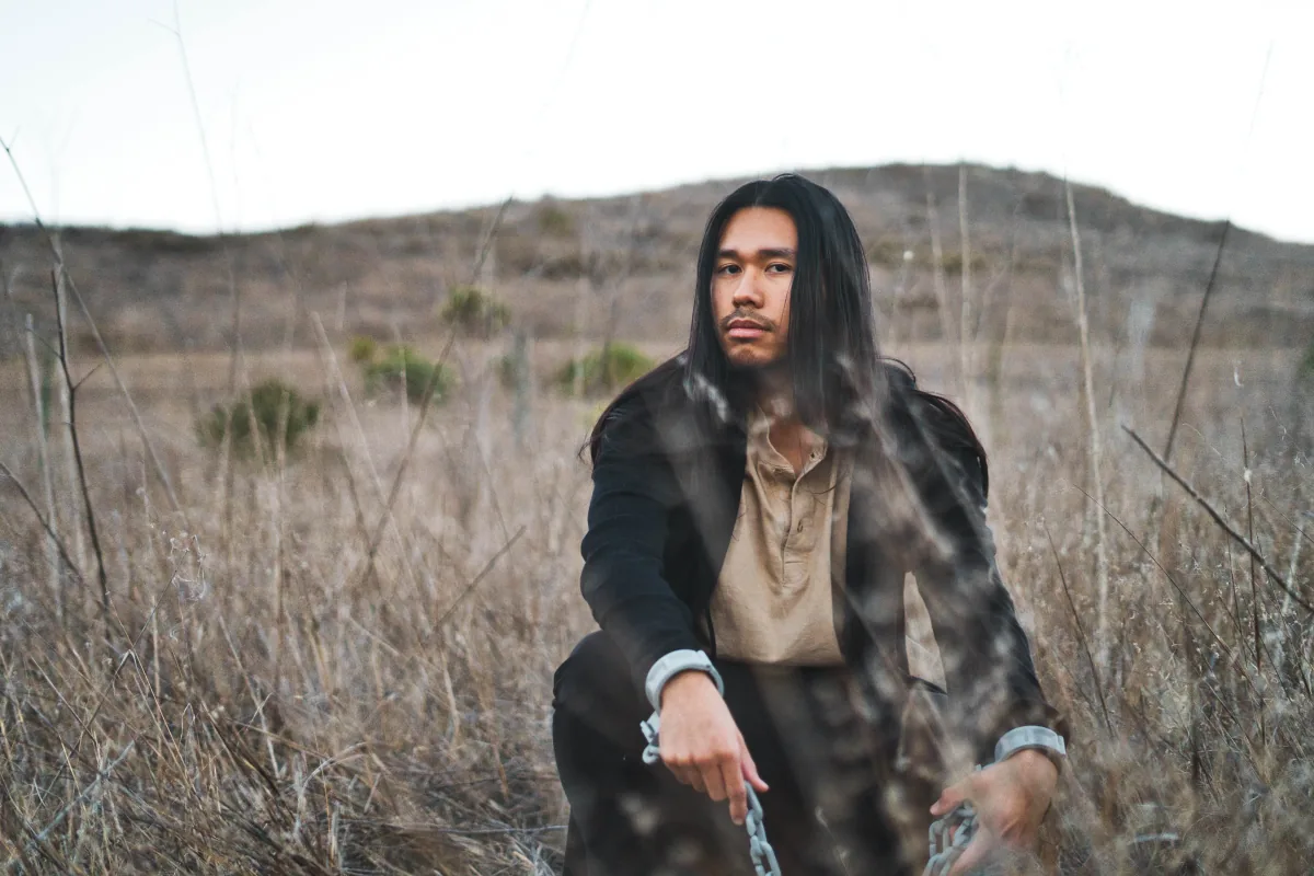 A man in a dark coat crouches in dry brush holding a chain, hillside behind him