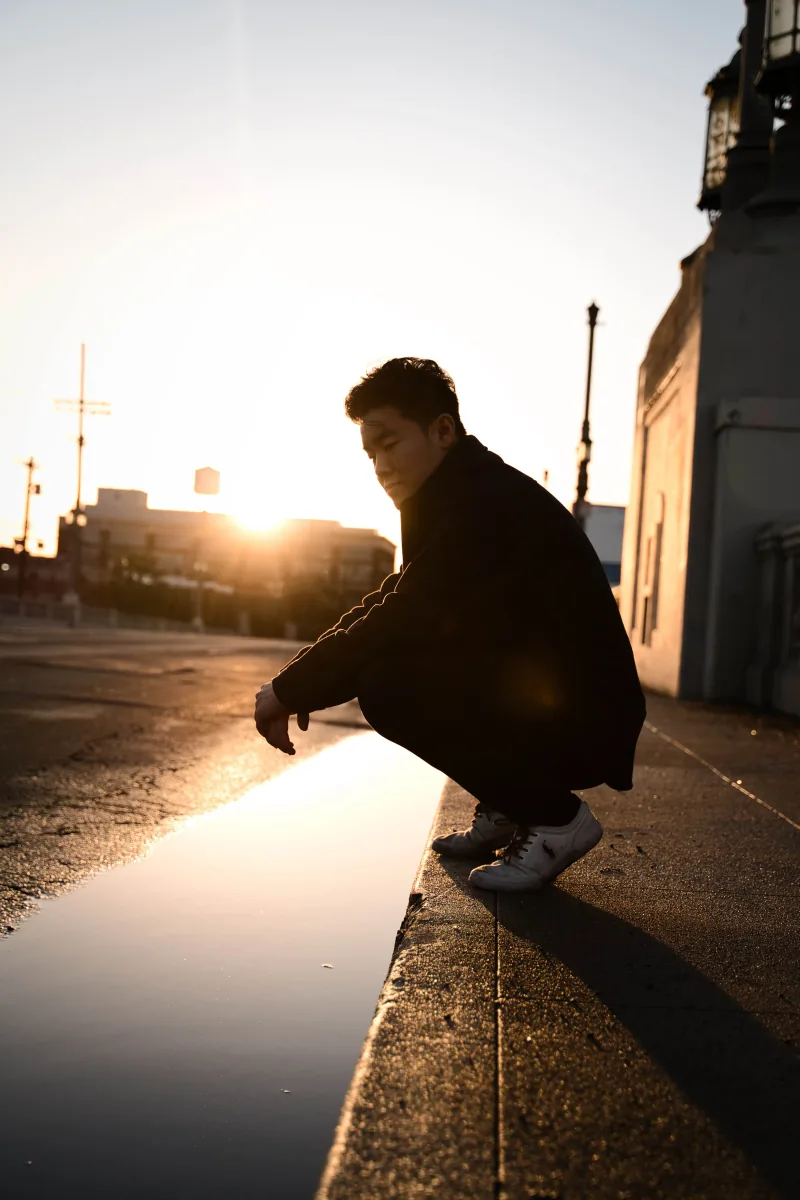 A man crouches on a curb in silhouette, the low sun flaring off the wet street beside him