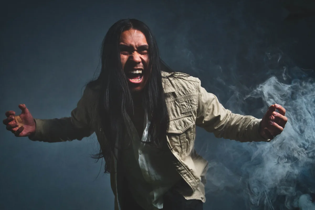 A man with long hair screams with arms flung wide, smoke drifting through the studio light