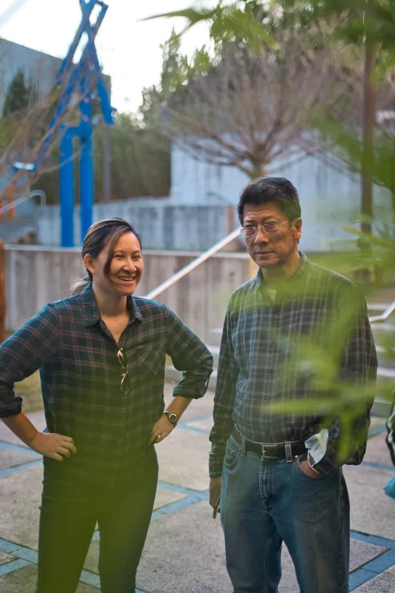 A woman laughing beside an older man, both in plaid, seen through out-of-focus leaves