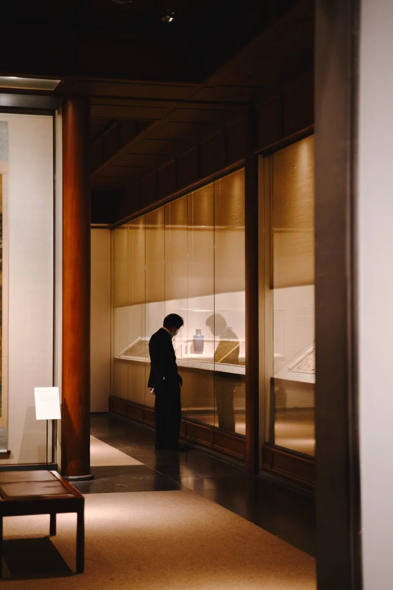 A lone visitor bends to read a museum vitrine in a dim wood-panelled gallery