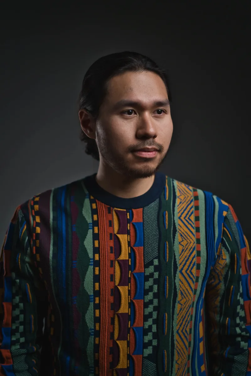 Studio portrait against near-black, a man in a boldly patterned knit sweater