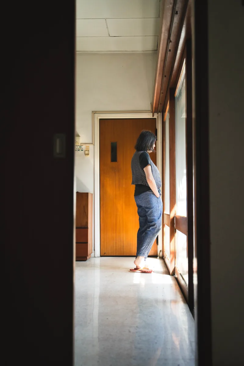 A woman stands at a sunlit window at the end of a dark hallway, seen through a doorway