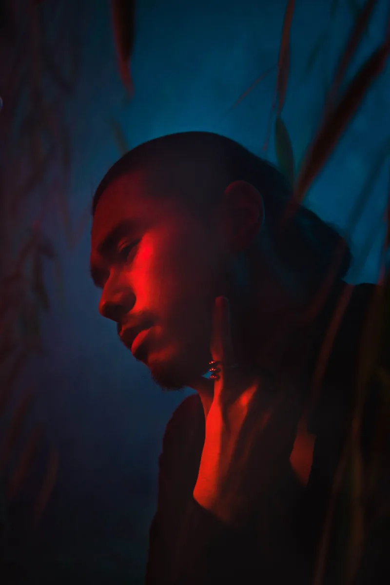 Profile lit deep red against blue darkness, hand raised to an earring, reeds crossing the frame