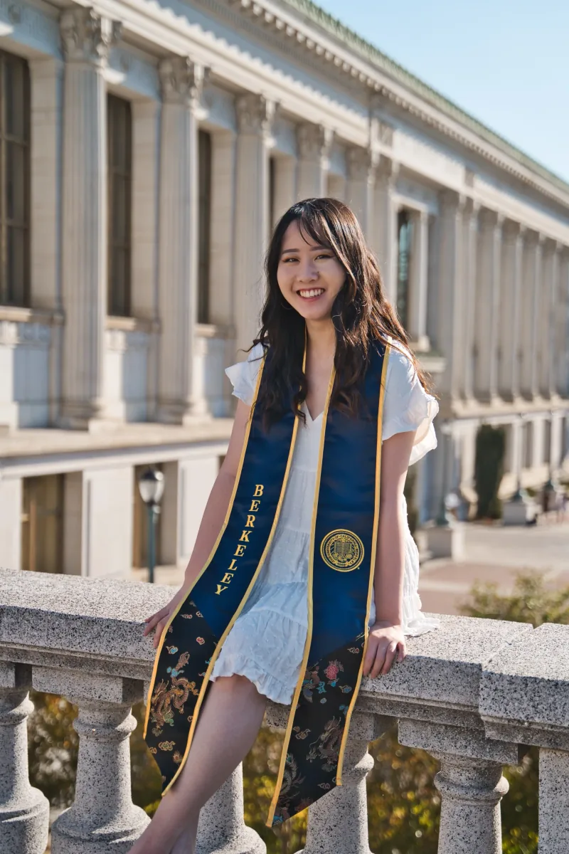 A graduate in a white dress and Berkeley stole laughs, perched on a stone balustrade outside Doe Library